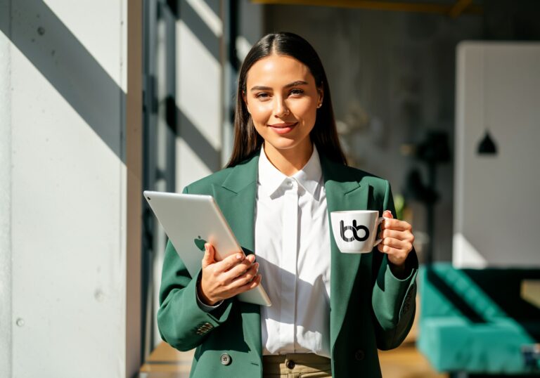 A woman in green blazer holding a mug with Baden Bower logo and a tablet smiling.