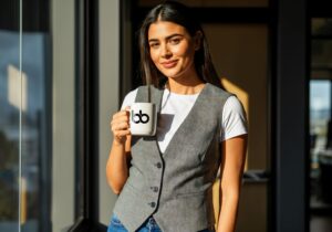 Woman in a gray vest and white tee standing by a window with a branded mug, exuding chic, interview-ready confidence, get featured in Forbes.