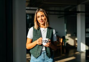 Professional in a green plaid vest and white tee holding a branded mug in a bright workspace, projecting a polished, magazine-ready look, get featured in Forbes.